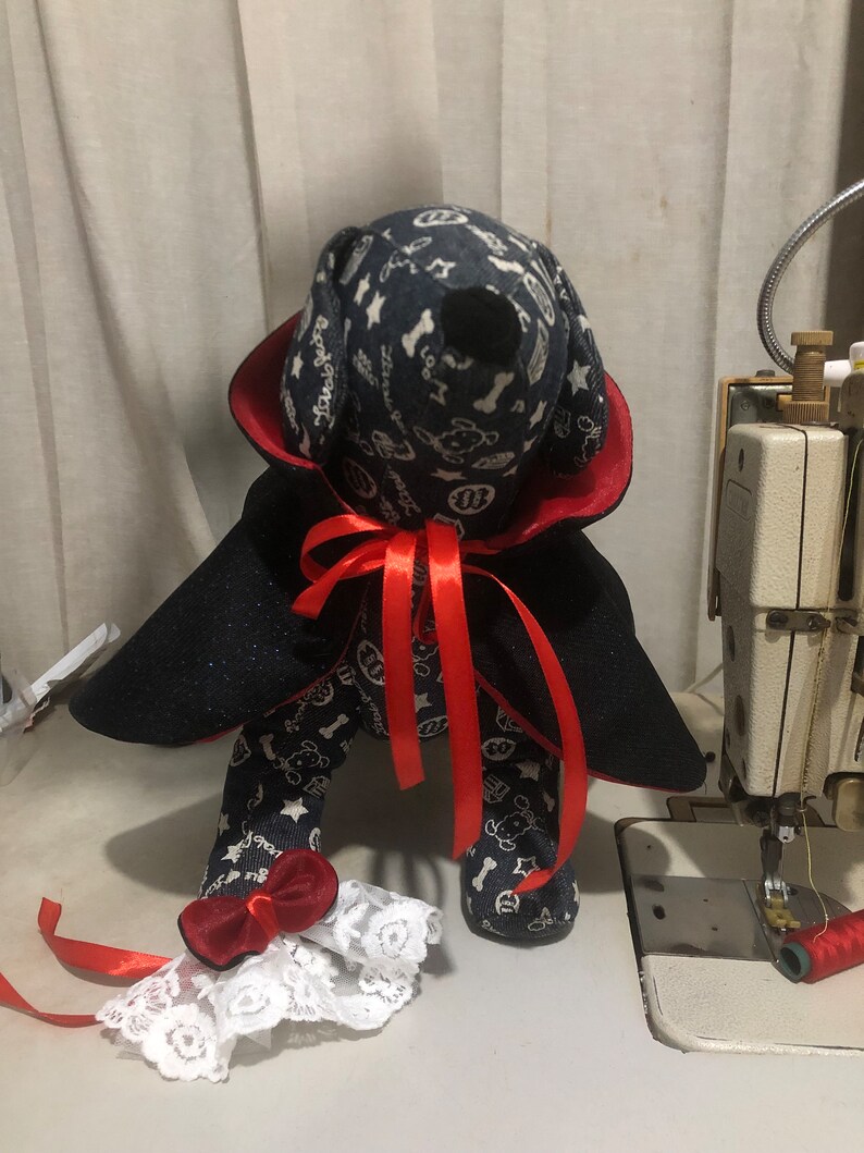 Puede incluir: Un perro de peluche negro y rojo vestido de vampiro con una capa negra, un collar rojo y una cinta roja. El perro lleva un brazalete de encaje blanco con un lazo rojo en cada pierna.