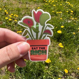 Puede incluir: Una pegatina con una Venus atrapamoscas en una maceta de terracota. La Venus atrapamoscas tiene hojas rojas y verdes. La maceta tiene el texto "EAT THE RICH" en verde y blanco. La pegatina se sostiene sobre un fondo de hierba verde y flores amarillas.