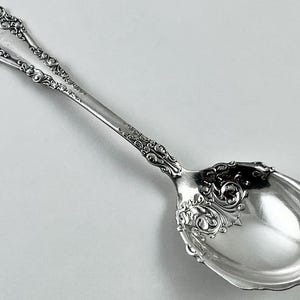 May include: A silver spoon with ornate, scrolling floral and foliate designs. The spoon features a long handle with a split design at the top and a rounded bowl. The spoon is likely made of sterling silver or a similar metal.