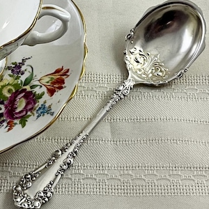 May include: A silver-toned metal spoon featuring an ornate, embossed scrollwork pattern along the handle and the base of the bowl. The spoon is positioned next to a white porcelain teacup decorated with a colorful floral pattern and gold trim.