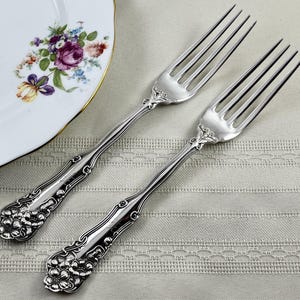 May include: Two silver-toned metal forks featuring ornate floral relief detailing on the handles. The forks have four tines and a polished finish, with intricate embossed flower patterns extending from the base of the handle to the neck.
