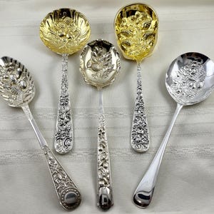 May include: A collection of five decorative metal spoons featuring intricate embossed designs of fruits, flowers, and foliage. Two spoons have a gold-toned finish, while three have a silver-toned finish. Each handle displays detailed relief patterns.