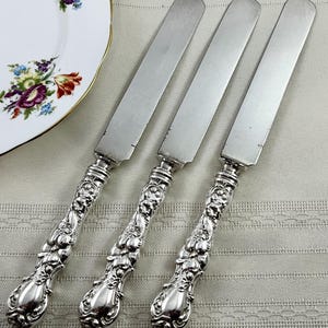 May include: Three silver-toned metal knives featuring smooth, flat blades and ornate, embossed handles with intricate floral and scrollwork detailing. The knives are arranged side-by-side, showcasing a polished metallic finish and decorative metalwork.