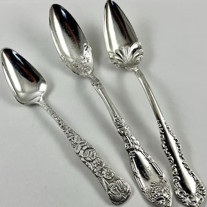 May include: Three antique silver spoons with ornate floral and vine detailing. The spoons have different bowl shapes and handle designs, showcasing intricate craftsmanship. The spoons are displayed on a white surface, highlighting their polished finish.