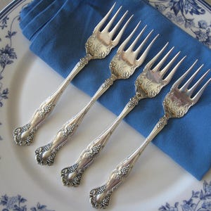 May include: Four ornate silver forks with detailed floral patterns are arranged on a white plate with a blue floral border. A folded blue napkin provides a backdrop for the forks, creating a classic and elegant table setting.