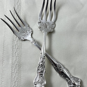 May include: Two silver forks with ornate floral and grape vine detailing. The forks are crossed, showcasing the intricate design on the handles and tines. The silverware is set against a neutral background, highlighting the elegant craftsmanship.