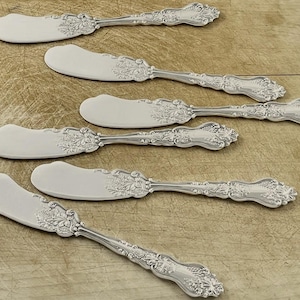May include: Six silver butter knives with ornate floral and leaf designs on the handles and blades. The knives are arranged on a light brown wooden surface, showcasing their elegant details.
