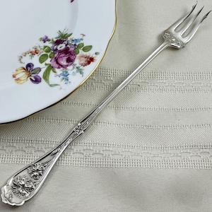 May include: A silver-toned metal fork with three tines and an elongated handle featuring embossed floral detailing at the base and near the tines. The fork is positioned diagonally next to a white ceramic plate with a colorful floral pattern.