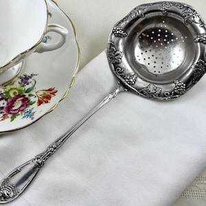 May include: A silver-toned metal tea strainer with a long handle and a circular bowl featuring a perforated center. The rim of the strainer and the handle are decorated with a raised relief pattern of grape clusters and vine leaves.