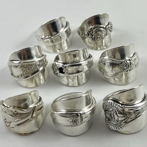 May include: A collection of eight silver-toned rings crafted from repurposed metal spoons. Each ring features a unique embossed pattern, including floral motifs, scrollwork, and geometric designs, with the curved handle forming the band.