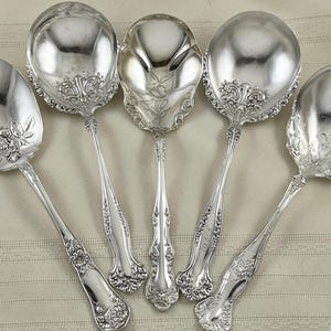 May include: Five ornate silver serving spoons with detailed floral and scroll designs. The spoons have large, rounded bowls and long, slender handles. The spoons are arranged in a fan shape on a light-colored surface.