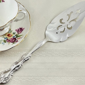 May include: A silver-toned metal cake server featuring an ornate, embossed handle with scrollwork details and a triangular blade with a decorative, symmetrical cutout pattern, positioned next to a white porcelain teacup with floral motifs.