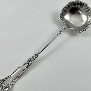 May include: A silver ladle with an ornate handle and a scalloped bowl. The handle features detailed scrollwork and a decorative design. The ladle is made of a shiny, reflective metal, likely silver, and is designed for serving.