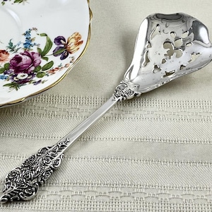 May include: A silver-toned serving spoon featuring an ornate, embossed floral handle and a wide, heart-shaped bowl with intricate cut-out detailing. The metal surface is polished and reflective, showcasing detailed craftsmanship on the handle.