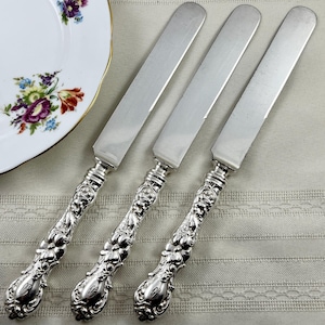 May include: Three silver-plated dinner knives featuring ornate, embossed floral handles with a polished finish. The knives have smooth, flat blades and detailed, decorative grips that taper toward the base, showcasing intricate metalwork.