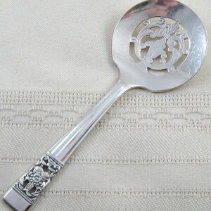 May include: A silver-toned serving spoon with a wide, circular bowl featuring a central cut-out scrollwork design. The handle is straight with a ribbed center and terminates in a decorative floral relief pattern at the base of the stem.