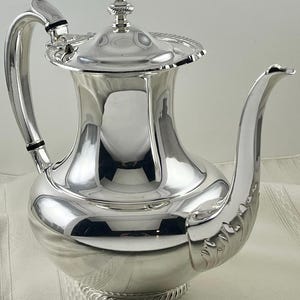 May include: A polished silver coffee pot with a curved spout and handle. The pot has a decorative lid with a finial and a rope-like design around the base. The pot is reflective, showing the surrounding environment.