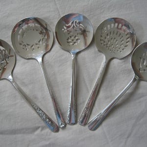 May include: Five silver serving spoons with decorative pierced designs. Each spoon has a different pattern on the bowl, including floral and geometric shapes. The handles have embossed floral detailing. The spoons are arranged on a light-colored surface.