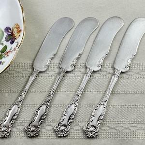 May include: Four silver-toned butter knives with rounded, wide blades and ornate, embossed scrollwork detailing on the handles. The knives are arranged side-by-side, showcasing a polished metallic finish and a consistent decorative pattern.