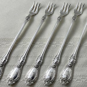 May include: A set of five silver-toned metal cocktail forks featuring long, slender handles with ornate floral embossing at the base and a monogrammed center. Each fork has three tines and a polished finish, arranged in a parallel row for display.