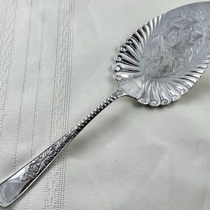 May include: Silver pie server with a decorative leaf design on the blade. The handle features an ornate pattern and a beaded section. The server has a scalloped edge and is designed for serving pies and cakes.