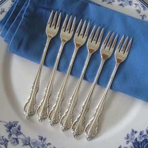 May include: Six silver forks with ornate handles are arranged on a white plate with a blue floral design. A folded blue napkin rests beneath the forks. The forks are placed on the plate, ready for a meal.