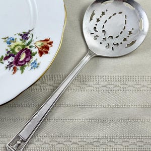 May include: A silver-toned slotted serving spoon with a circular bowl featuring intricate cut-out patterns. The handle is straight with a tapered design and a decorative rectangular motif at the base, showcasing a classic metallic finish.