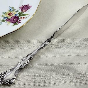 May include: A silver-toned metal butter knife featuring an ornate, embossed handle with intricate scrollwork and leaf-like detailing. The blade is smooth and slightly curved, extending from the decorative handle to a pointed tip.