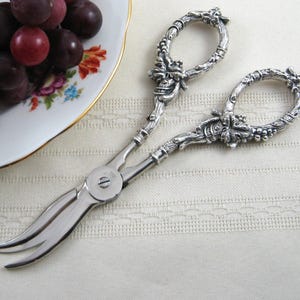 May include: Silver-toned metal grape shears featuring ornate, textured handles with detailed vine and leaf motifs. The blades are smooth and curved, designed for cutting fruit. The handles are shaped like twisted branches with clusters of grapes.