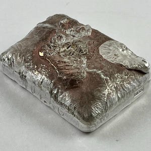 May include: A rectangular silver bar with a textured, oxidized copper-toned center surface. The edges are polished silver, and the top features raised, organic, molten-style silver relief details. The side is stamped with the text 104.5G.