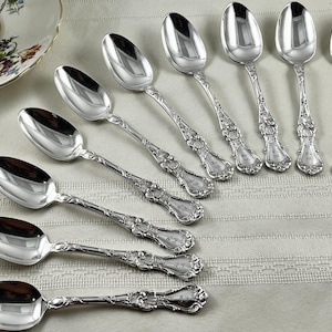 May include: A collection of silver spoons with ornate floral detailing on the handles. The spoons have a reflective, polished surface, and are arranged on a light-colored, textured surface. A floral teacup and saucer are visible in the upper left corner.