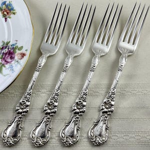 May include: Four silver-toned metal forks featuring ornate floral relief detailing on the handles. Each handle includes a decorative scrollwork pattern and a central engraved monogram. The forks have four tines and a polished, reflective finish.
