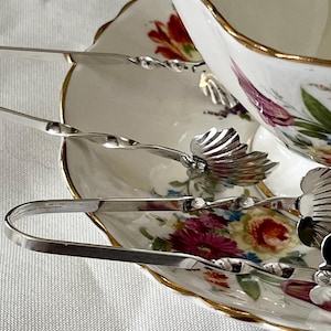 May include: Silver sugar tongs with a seashell design rest on a floral patterned saucer next to a teacup with a gold rim. The teacup and saucer have a white background with red, pink, and yellow flowers.
