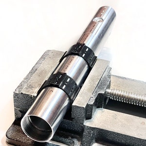 Pode incluir: Um tubo de metal prateado com bra&ccedil;adeiras de pl&aacute;stico preto est&aacute; preso em um torno de metal. O torno tem uma al&ccedil;a rosqueada.