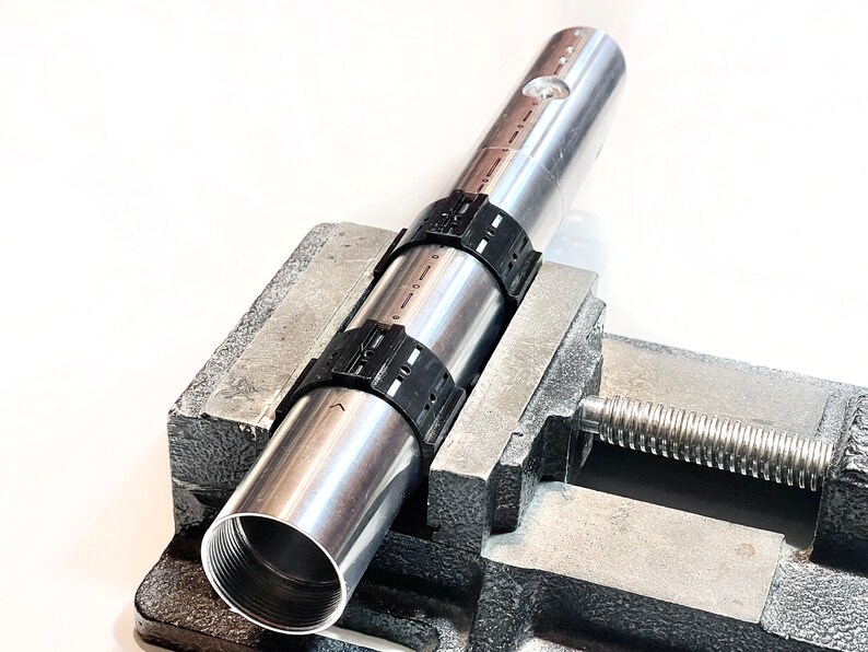 Pode incluir: Um tubo de metal prateado com bra&ccedil;adeiras de pl&aacute;stico preto est&aacute; preso em um torno de metal. O torno tem uma al&ccedil;a rosqueada.
