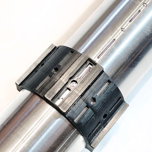 Pode incluir: Close-up de uma bra&ccedil;adeira de metal preta e prateada com uma s&eacute;rie de ranhuras e furos. A bra&ccedil;adeira est&aacute; presa a um tubo de metal prateado.
