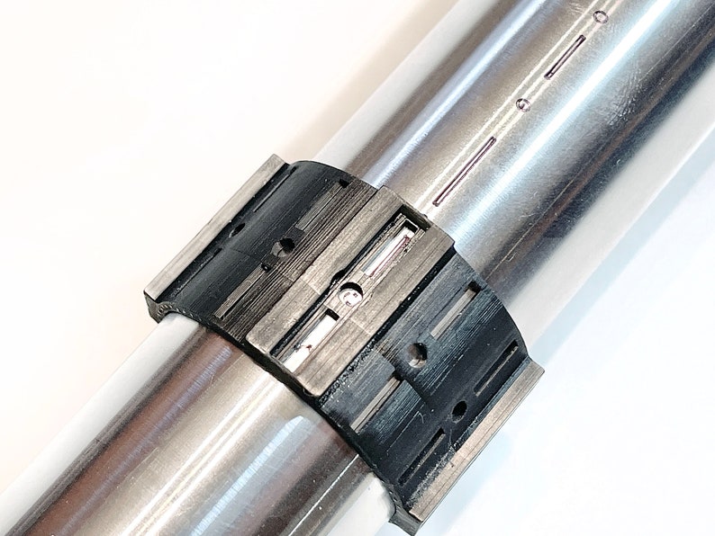 Pode incluir: Close-up de uma bra&ccedil;adeira de metal preta e prateada com uma s&eacute;rie de ranhuras e furos. A bra&ccedil;adeira est&aacute; presa a um tubo de metal prateado.