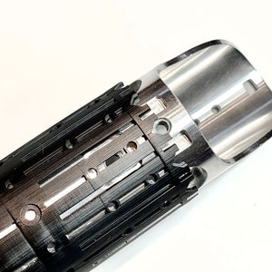 Pode incluir: Um close-up de um objeto de metal preto e prata com uma s&eacute;rie de pequenos orif&iacute;cios e ranhuras. O objeto &eacute; cil&iacute;ndrico e tem uma extremidade arredondada.