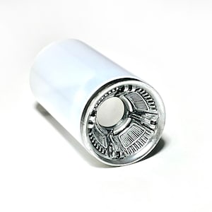 Puede incluir: Objeto cilíndrico de metal blanco con un interior detallado de color plateado. El interior presenta un diseño geométrico complejo. El objeto está sobre un fondo blanco.