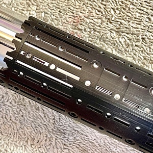 Pode incluir: Uma empunhadura de sabre de luz preta com uma superf&iacute;cie texturizada e v&aacute;rios orif&iacute;cios circulares. A empunhadura tem uma se&ccedil;&atilde;o prateada na parte superior.