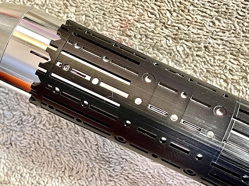 Pode incluir: Uma empunhadura de sabre de luz preta com uma superf&iacute;cie texturizada e v&aacute;rios orif&iacute;cios circulares. A empunhadura tem uma se&ccedil;&atilde;o prateada na parte superior.