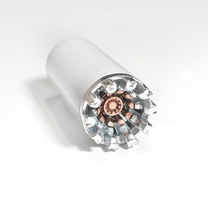 Puede incluir: Un objeto metálico de color plateado y cobre con un cuerpo cilíndrico y un extremo en forma de estrella. El objeto tiene un fondo blanco.