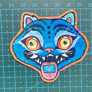 May include: A vibrant sticker of a cartoon cat's face, with blue fur, an orange border, and large yellow eyes. The cat has a pink nose, white teeth, and a tongue with a small image. The sticker has a glittery effect.