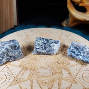 May include: Three blue-grey tumbled stones on a wooden surface with a circular design. The stones are rough and have a natural, unpolished look.