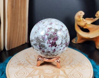May include: A white and pink speckled crystal sphere on a copper stand. The sphere is sitting on a wooden base with a circular design.
