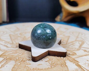 May include: A dark green orb with white speckles rests on a small, light-colored wooden star. The orb is smooth and reflective, and the star-shaped base has a dark brown edge. The background is blurred, with hints of a wooden surface.