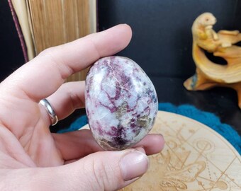 May include: A smooth, polished, pink and white egg-shaped crystal with a swirling pattern. The crystal is being held in a hand.