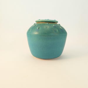 May include: A teal ceramic vase with a rounded body and a small, flat lid. The vase has a textured surface with small, raised dots. The lid is a lighter shade of teal and has similar dot detailing. The vase is a decorative item.