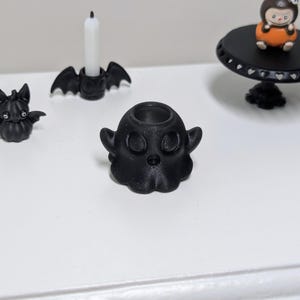 Peut inclure: Décorations noires sur le thème d'Halloween, dont un bougeoir en forme de fantôme, une figurine de chauve-souris et un bougeoir avec des ailes de chauve-souris. Une petite table avec une figurine est également visible.