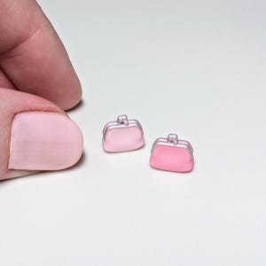 Op de afbeelding: Twee miniatuur roze portemonnees met zilverkleurige accenten. De portemonnees zijn klein, met een afgeronde vorm en een sluitingsdetail. De afbeelding is een close-up, die de portemonnees in detail toont. De portemonnees liggen op een wit oppervlak.
